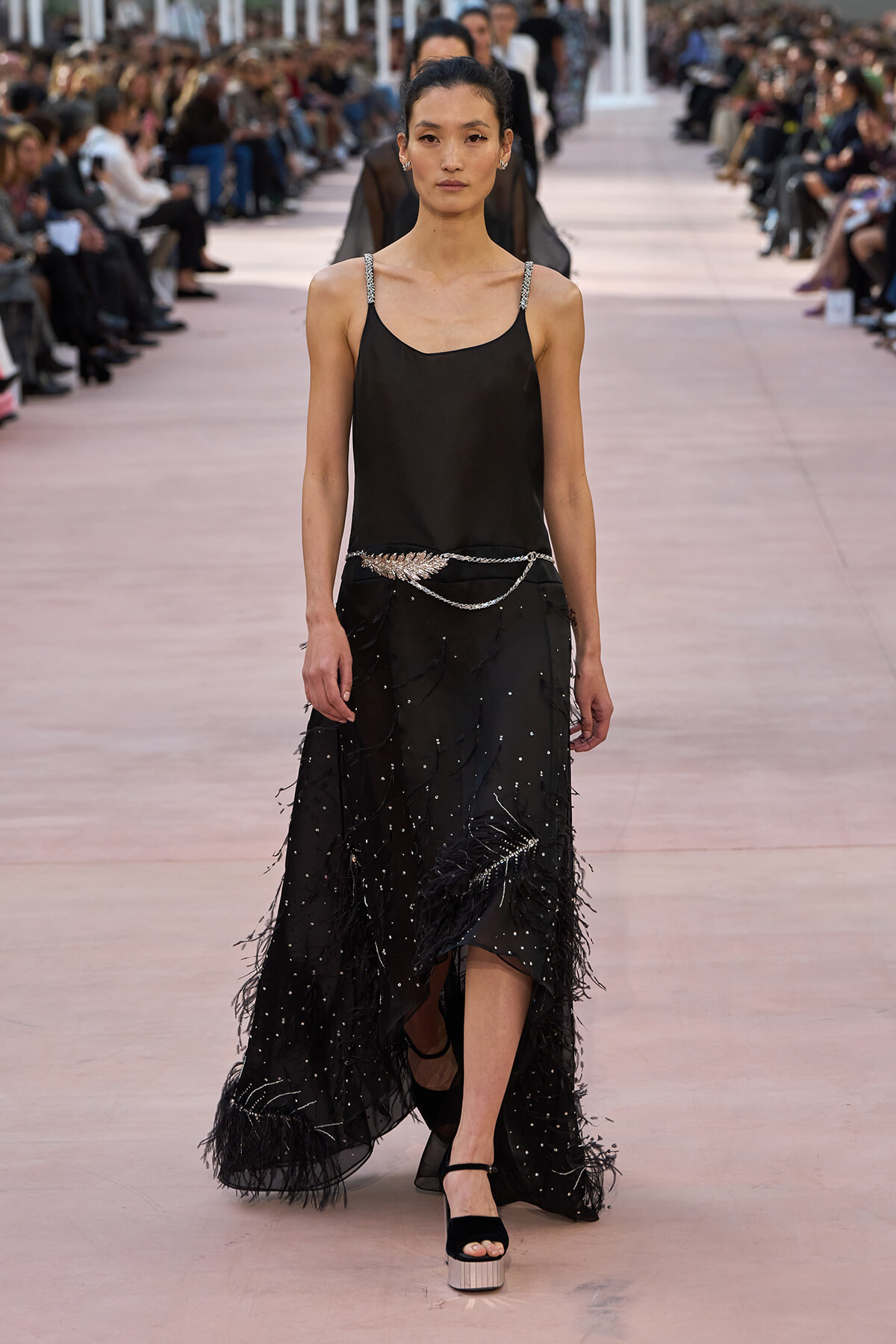 Model walks the runway in a black spaghetti-strap gown with a silver beaded belt and feathered hem.