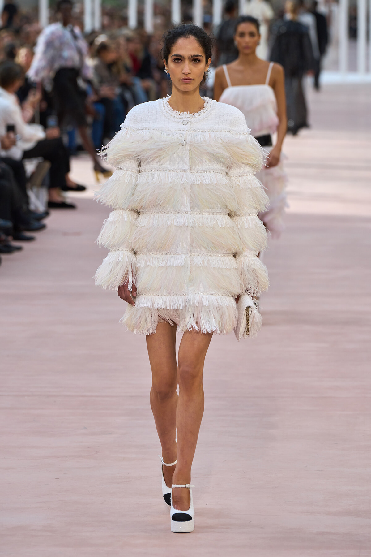 Model walking a runway in a white, layered feathered mini dress, holding a small clutch.