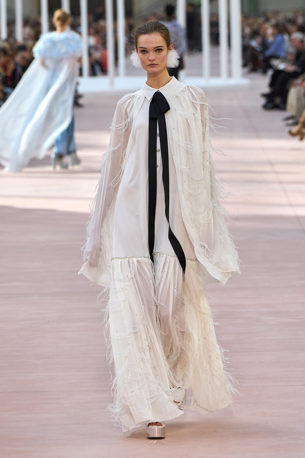 Model walks the runway in a white feathered, floor-length gown with a black ribbon tie at the neck.