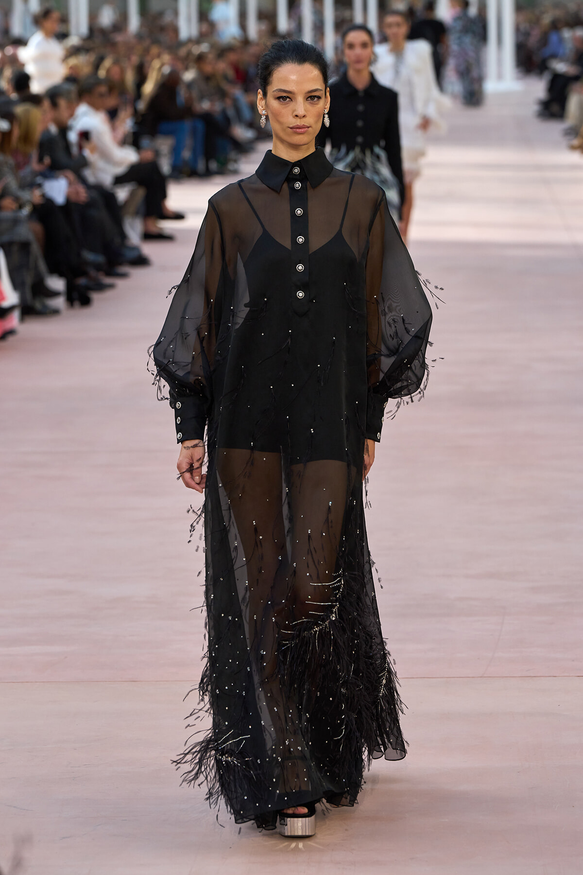 Runway model in a sheer black dress with silver buttons and feathered hem, walking the catwalk as audience watches.