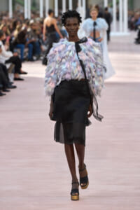 Model walking the runway in a pastel feathered top and black layered skirt, audience blurred in the background.