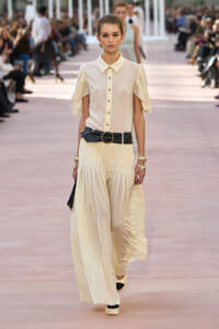 Model walking a runway in an ivory blouse with ruffled sleeves, cream wide-leg pleated pants, and a wide black belt with gold buckle.