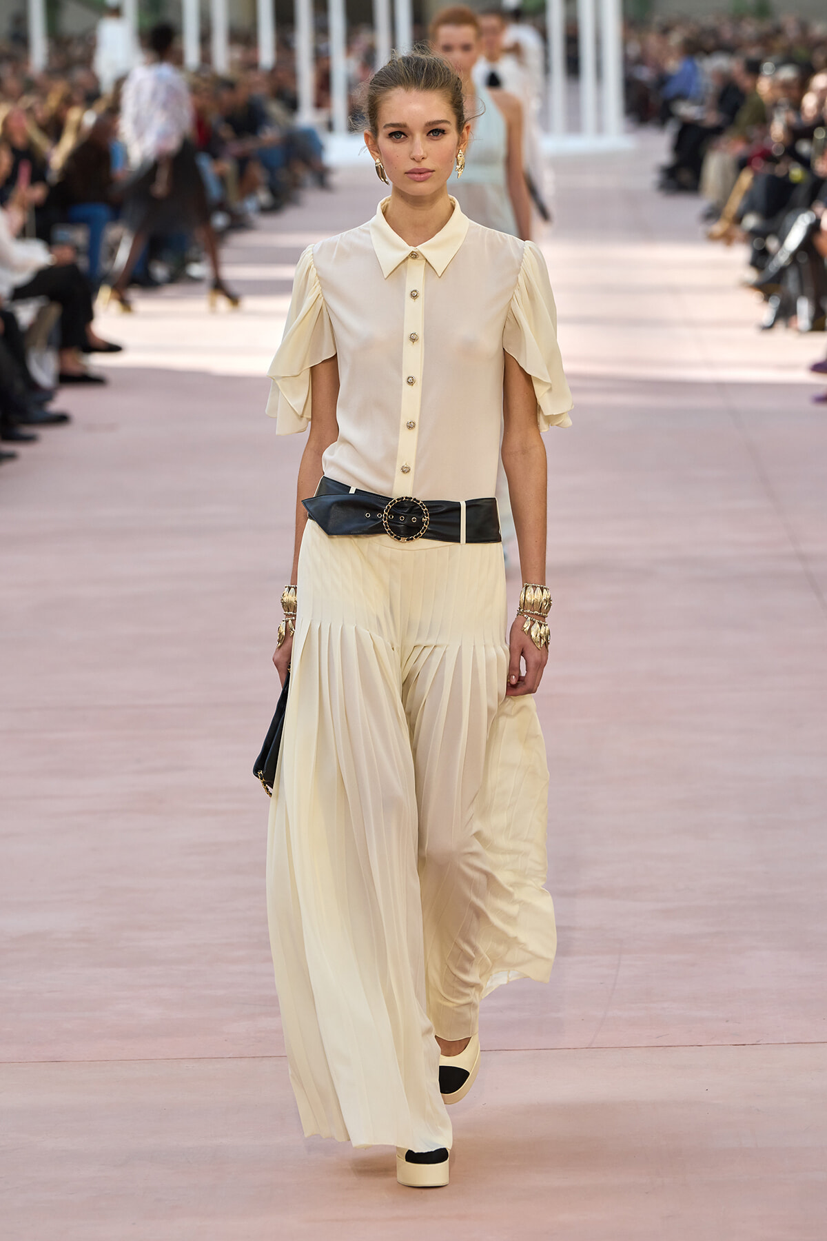 Model walking a runway in an ivory blouse with ruffled sleeves, cream wide-leg pleated pants, and a wide black belt with gold buckle.
