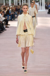 Model on a runway wearing a pale yellow pleated dress with cape-like overlay, gold belt, and matching heels, carrying a small yellow handbag.