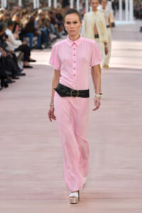 Model walking a runway in a pale pink jumpsuit with a wide black belt and gold accessories.