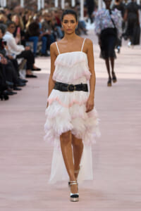 Model walking a runway in a white layered ruffled dress with pink accents, cinched by a wide black belt and metallic platform heels.