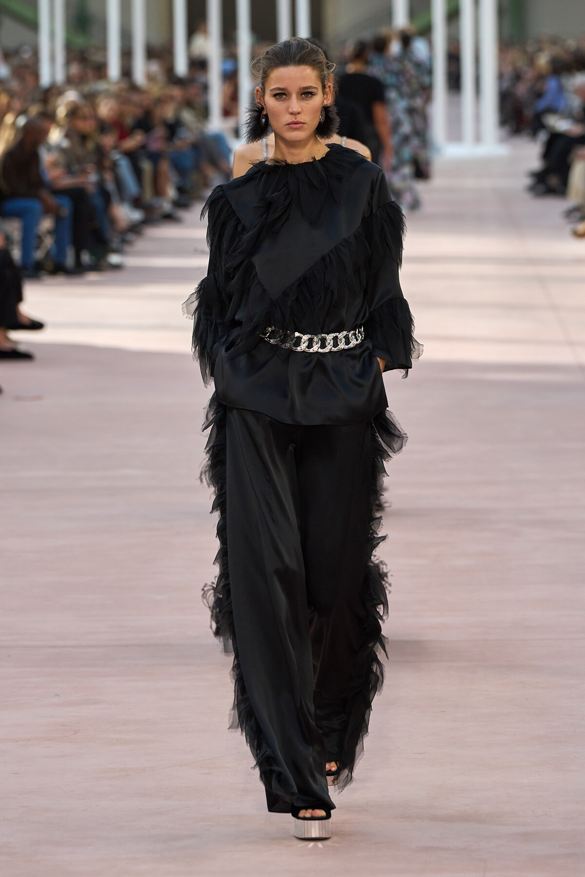 Model walks a runway wearing a black, ruffled, sheer-trim jumpsuit with a chunky silver chain belt and platform sandals.
