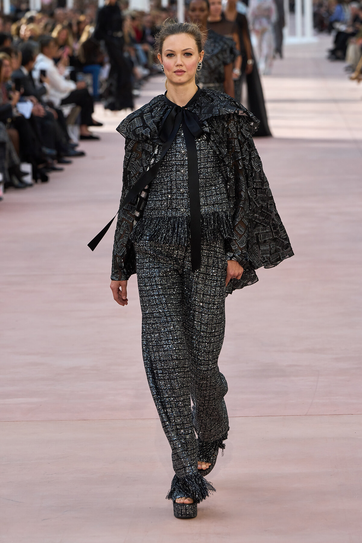 Model walks a fashion runway in a metallic tweed pantsuit with a layered cape, black bow at the neck, and platform sandals.