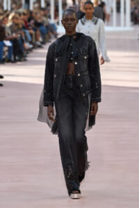 Model walks a runway in an all-black denim outfit: scarf-tied top, quilted denim jacket, sheer cape, and dark sequined jeans with fringe at the hem.