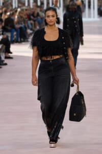 Model on a runway wearing a black fringe crop top, loose dark jeans with sequined cuffs, and a quilted black handbag with a gold chain.