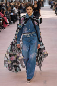 Model walking a fashion runway in a denim jumpsuit with embroidered flame-like patterns and a long sheer floral cape draped over shoulders.