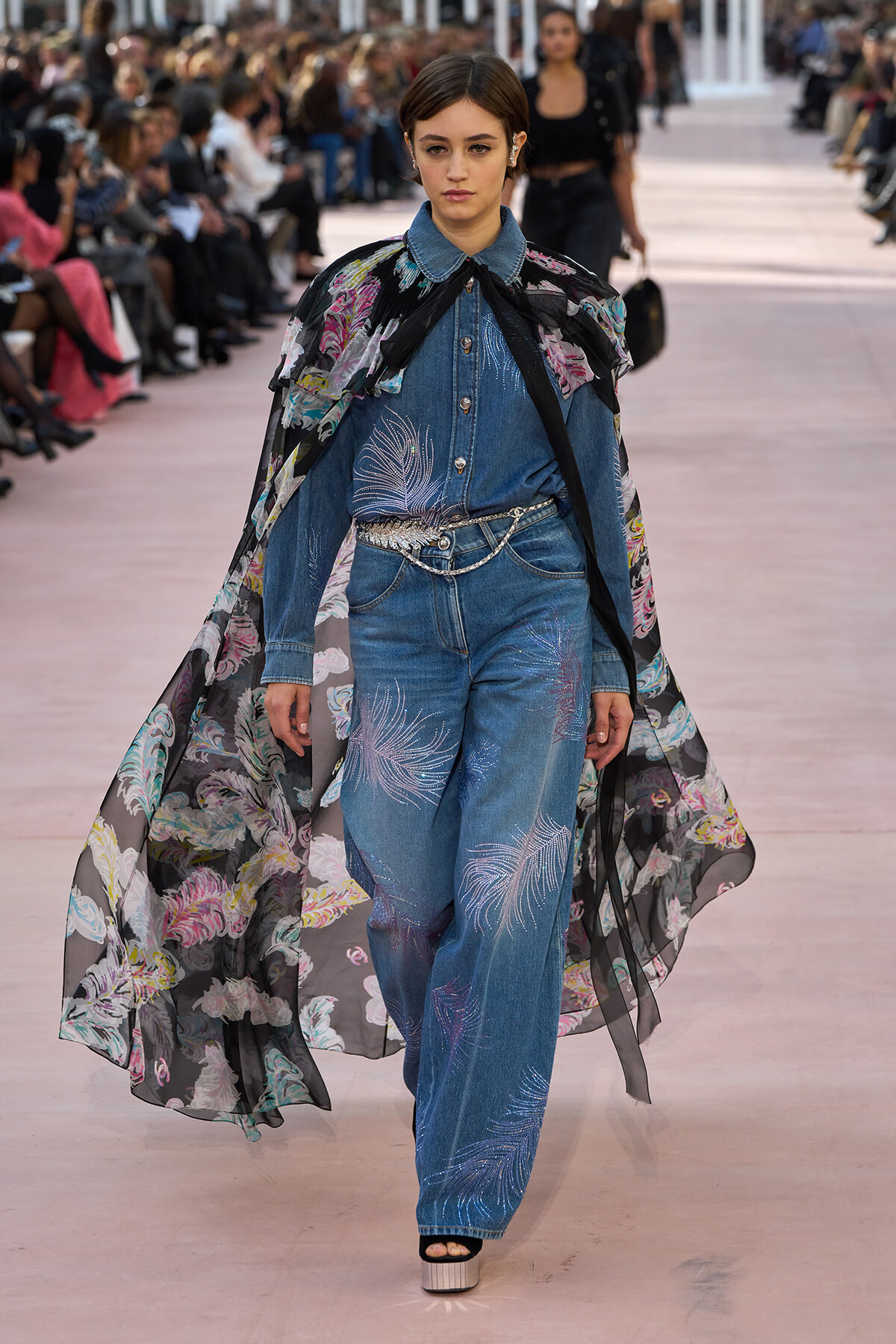 Model walking a fashion runway in a denim jumpsuit with embroidered flame-like patterns and a long sheer floral cape draped over shoulders.
