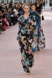Model walks the runway in a black floral-print jumpsuit with a ruffled collar and silver belt, carrying a glittery clutch. Audience watches from both sides.