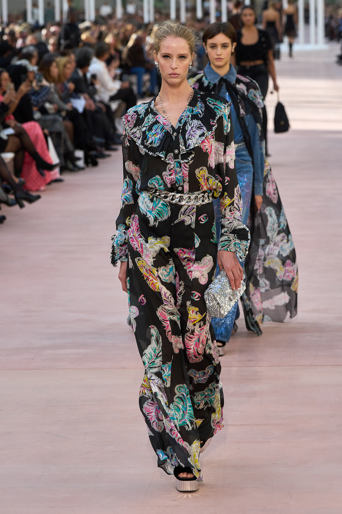 Model walks the runway in a black floral-print jumpsuit with a ruffled collar and silver belt, carrying a glittery clutch. Audience watches from both sides.