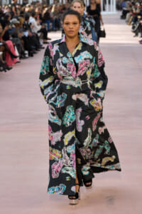 Model walking a fashion show in a long black coat with multicolored feather print and a silver belt.
