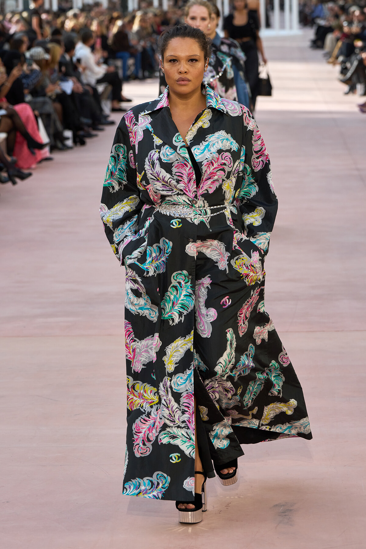 Model walking a fashion show in a long black coat with multicolored feather print and a silver belt.