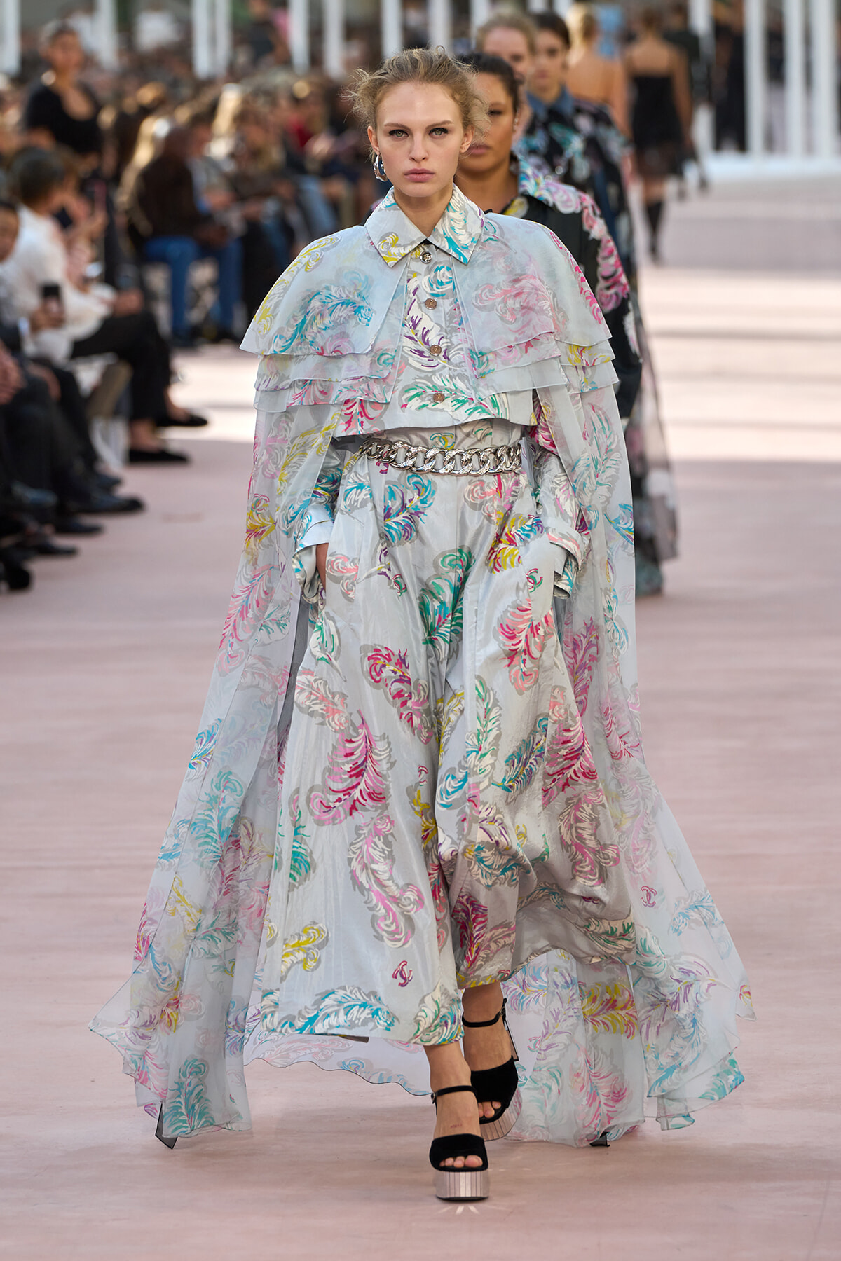 Model walks the runway in a pale blue floral cape and matching wide-leg outfit with layered ruffles and a chain belt.