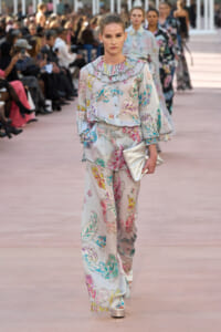 Model walks a pastel floral jumpsuit with ruffled collar and sleeves, carrying a metallic clutch on a fashion show runway with seated audience behind.