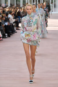 Model walking a fashion runway in a pastel floral romper with ruffled high neck and belt, audience blurred in background.