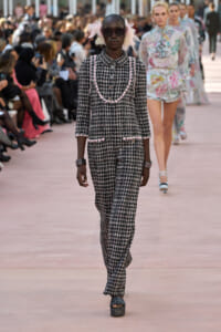Model walks the runway in a black-and-white plaid tweed suit with pink trim and accessories (pink feathers).