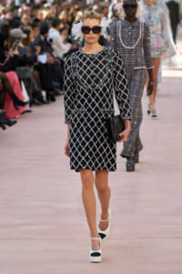 Runway model in a black-and-white lattice dress, oversized sunglasses, and white floral headpiece, carrying a black clutch.