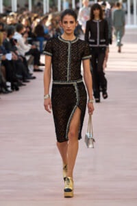 Model walks the runway in a black lace dress with silver trim and buttons, carrying a small metallic handbag and wearing gold platform shoes.