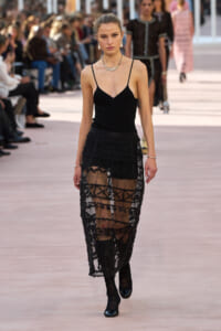 Model walking a runway in a black spaghetti-strap top and sheer lace skirt with opaque shorts underneath, audience in background