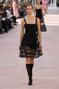 Model walks a fashion show in a black lace dress with a sheer hem, wearing jewelry, as audience watches from the sides.