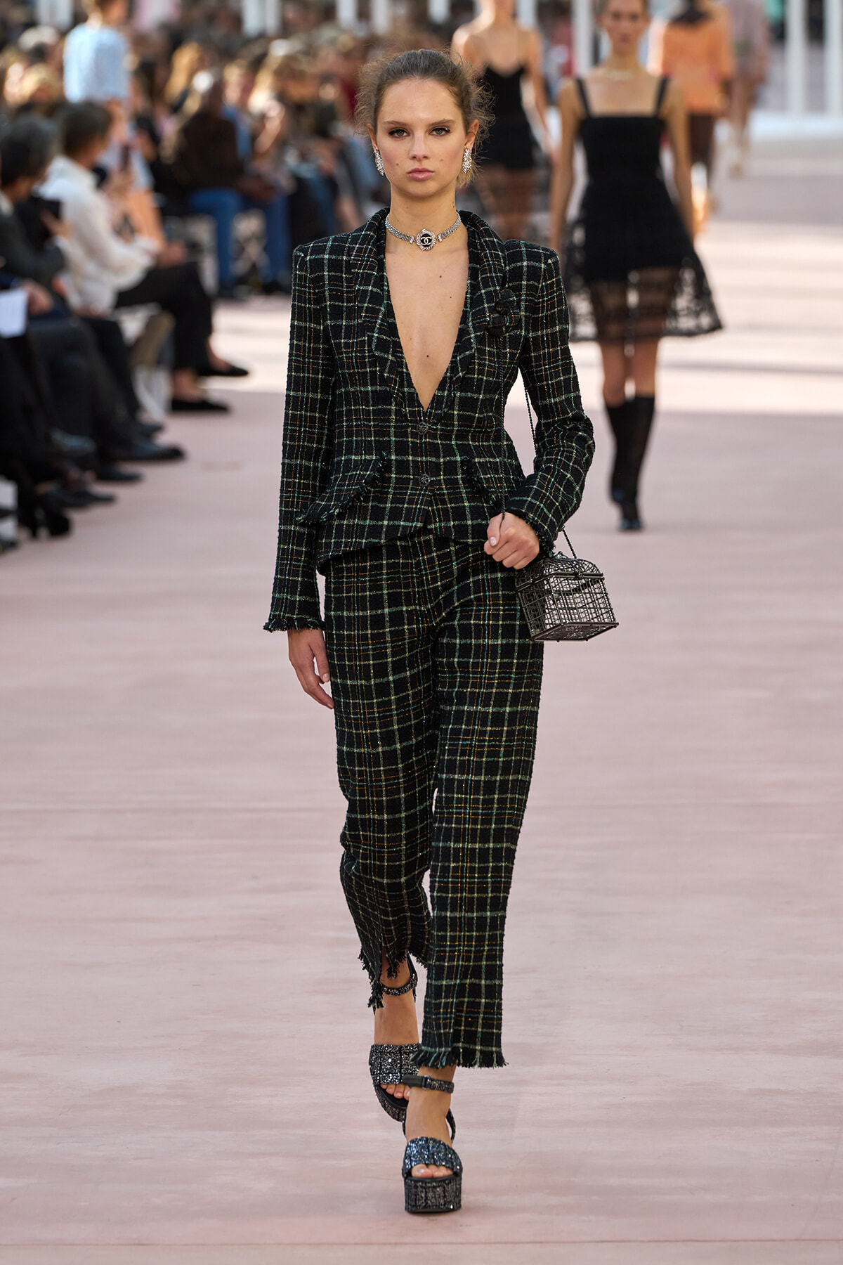 Runway model wearing a dark plaid tailored suit with a deep V neckline, silver jewelry, and a small lattice-style handbag