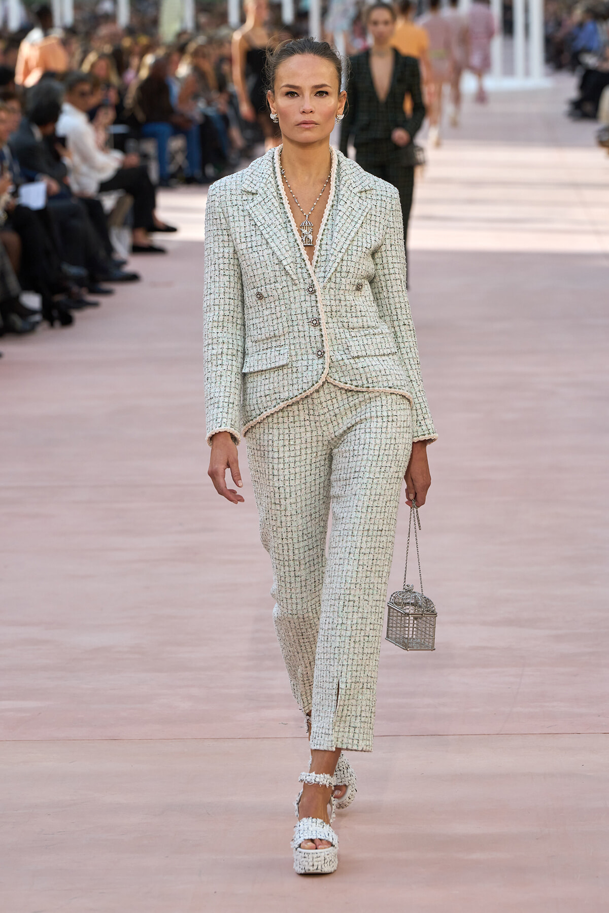Model walks the runway in a light tweed pantsuit with a matching blazer and pearl jewelry, carrying a small cage-style handbag.