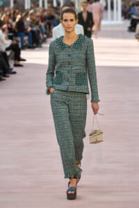 Model walks a fashion runway in a green plaid tweed suit with fringe detailing on the collar and pocket trims, carrying a small gold lattice handbag.
