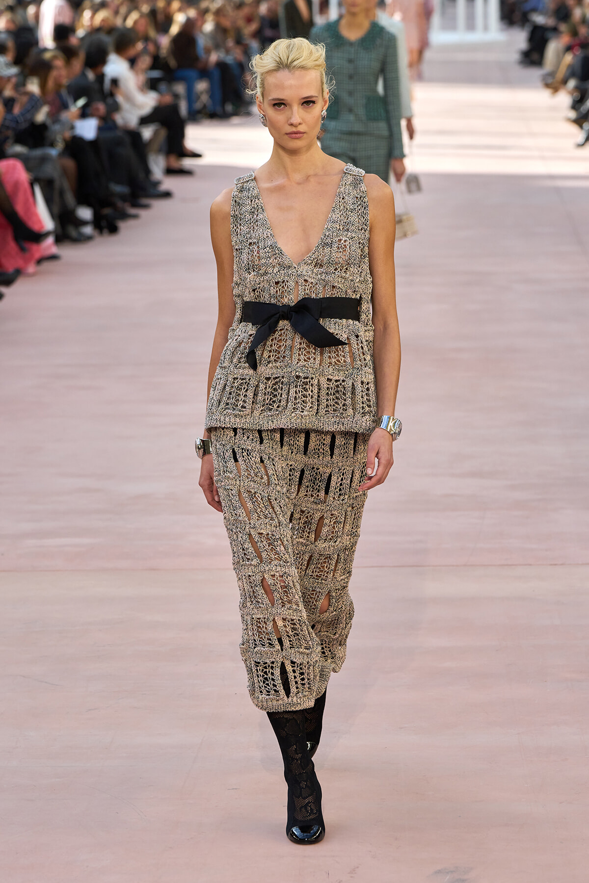 Model on a runway wearing a beige crocheted sleeveless dress with a black bow belt and a layered open-weave skirt, paired with black lace boots.