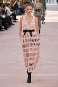 Model on a runway wearing a pale pink crochet maxi dress with a black bow belt, statement necklace, and black lace tights; audience blurred in the background.