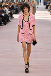 Model walking a runway in a pink textured dress with black trim, matching jacket, and black platform sandals; holding a black feathered clutch in the crowd-lit scene.