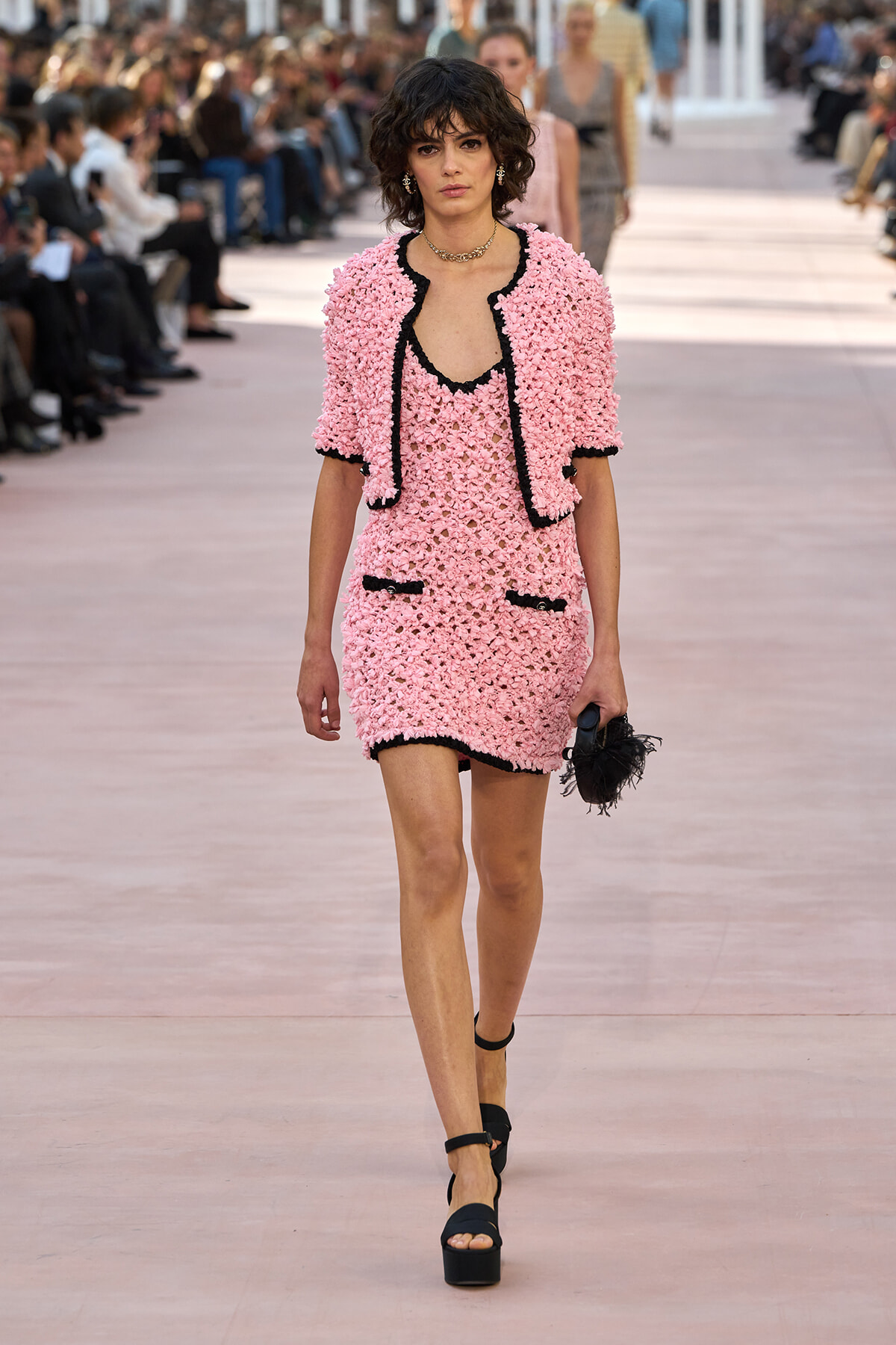 Model walking a runway in a pink textured dress with black trim, matching jacket, and black platform sandals; holding a black feathered clutch in the crowd-lit scene.