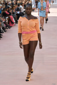Model walking a runway in a pink and yellow striped off-the-shoulder cardigan and matching shorts on a fashion show catwalk.