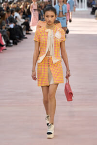 Model walks the runway in a pink-yellow tweed two-piece with a cream bow blouse, front-slit skirt, and pink handbag.