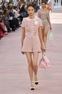 Model walking a fashion runway in a pink and lime tweed romper with a pink collar and large white flower accessory; carrying a pink quilted handbag and wearing strappy heels.