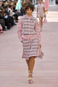 Model walking a pink plaid suit with puff sleeves down a runway at a fashion show.