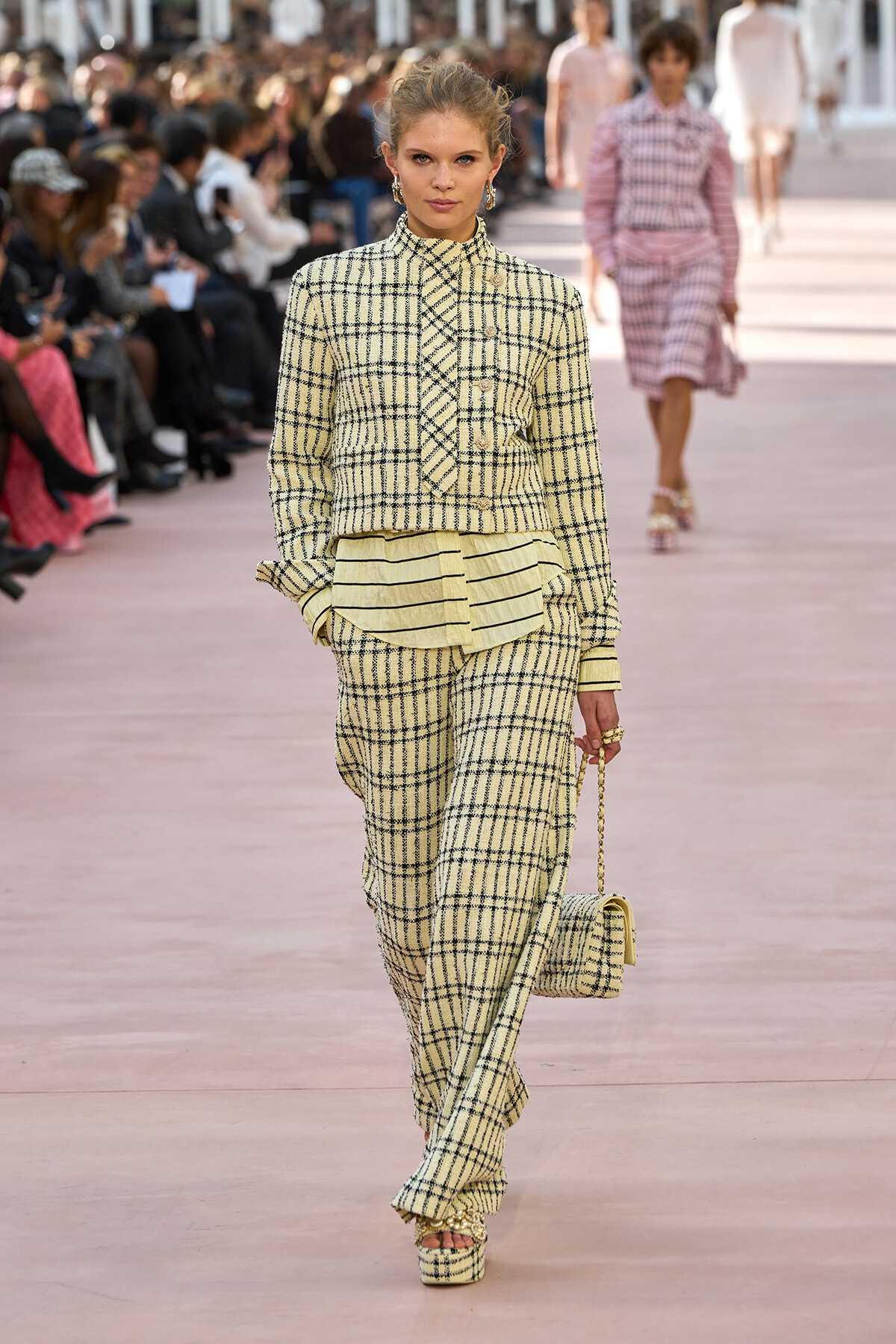 Model walks the runway in a cream plaid suit with a layered ruffled top, matching wide-leg pants, platform sandals, and a small plaid handbag.