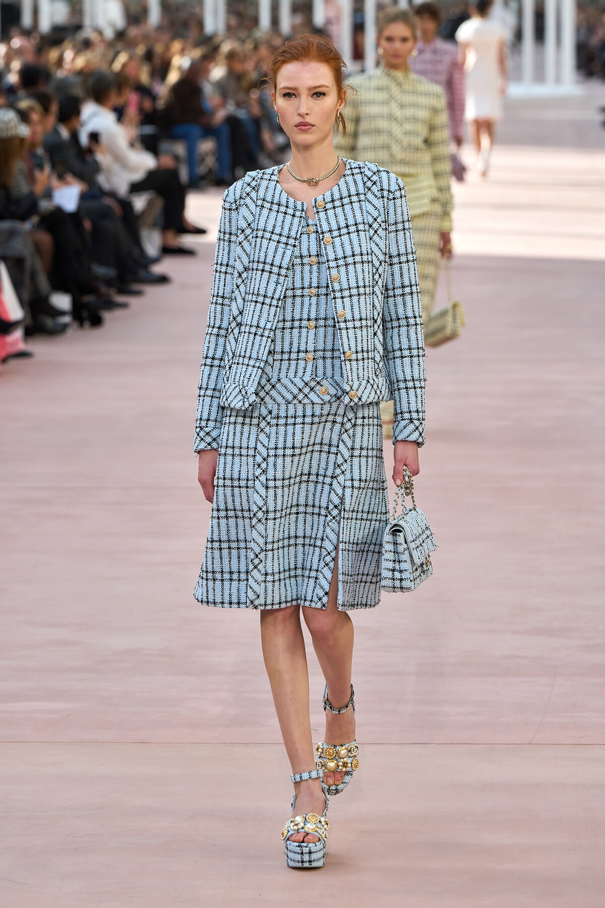 Model walking a runway in a light blue plaid matching jacket and knee-length skirt suit with gold buttons, carrying a small plaid handbag.