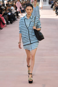 Model on a runway wearing a light blue striped cardigan over a matching striped outfit, carrying a black handbag and wearing embellished platform sandals.