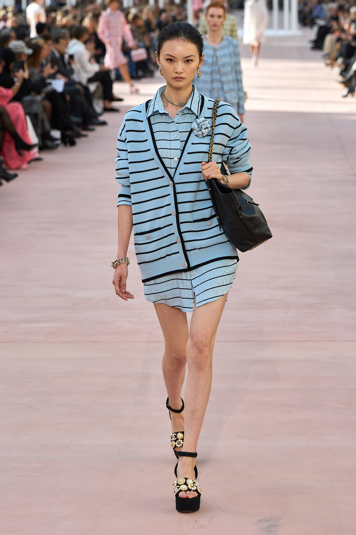 Model on a runway wearing a light blue striped cardigan over a matching striped outfit, carrying a black handbag and wearing embellished platform sandals.