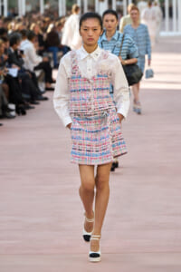 Model walks a fashion runway in a pastel plaid tweed dress over a white button-down blouse, with a floral embellishment at the chest.