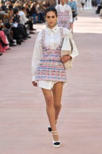 Model walking a fashion runway in a white shirt layered with a pastel plaid tweed mini-dress, carrying a white bag.
