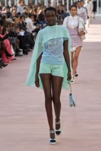 Model walks a fashion runway in a pastel mint cape over a light blue top and mint shorts, with a pearl necklace and pale blue handbag.