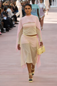 Model walks a fashion runway in a pale pink sheer cape over a beige matching top and skirt, carrying a pale yellow quilted bag.