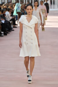 Model walking the runway in a white textured knee-length, double-breasted dress with ruffled cap sleeves.