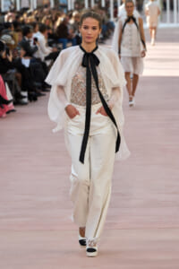 Model walking runway in white sheer cape over sequined top and wide white pants, black neck ribbon tie, audience in background