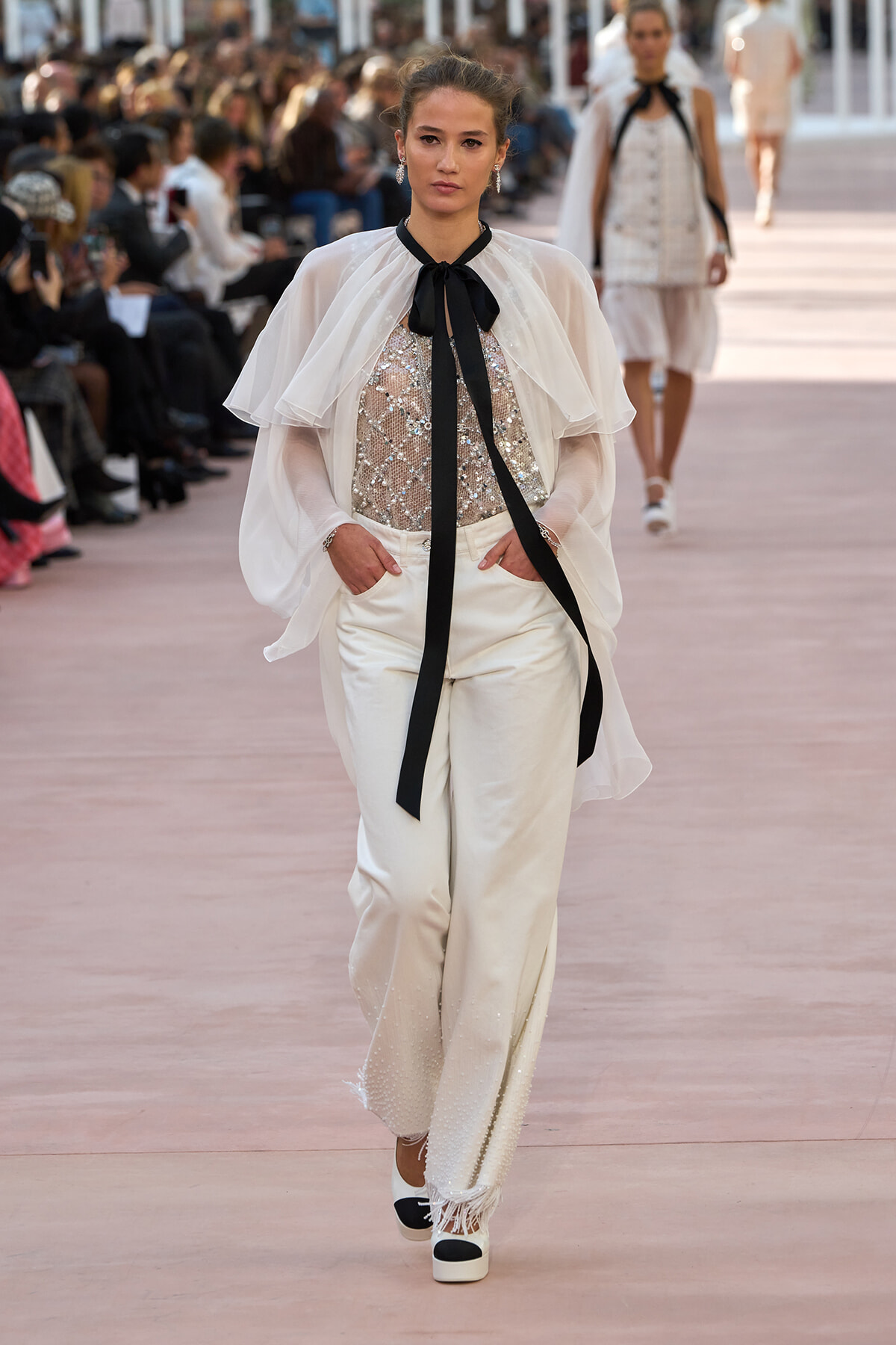 Model walking runway in white sheer cape over sequined top and wide white pants, black neck ribbon tie, audience in background